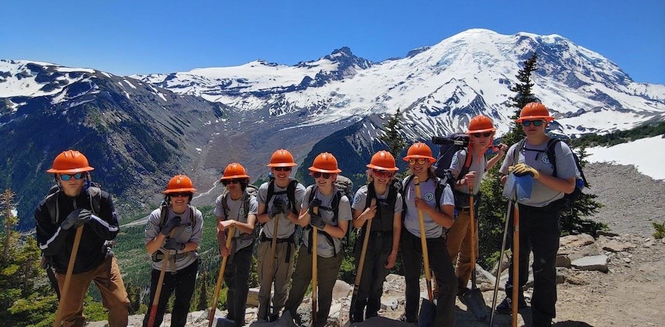 A row of young people wearing grey YCC shirts, orange hard hats, and holding shovels stand in front of a glaciated mountain peak with a glacier descending into a large valley behind them.