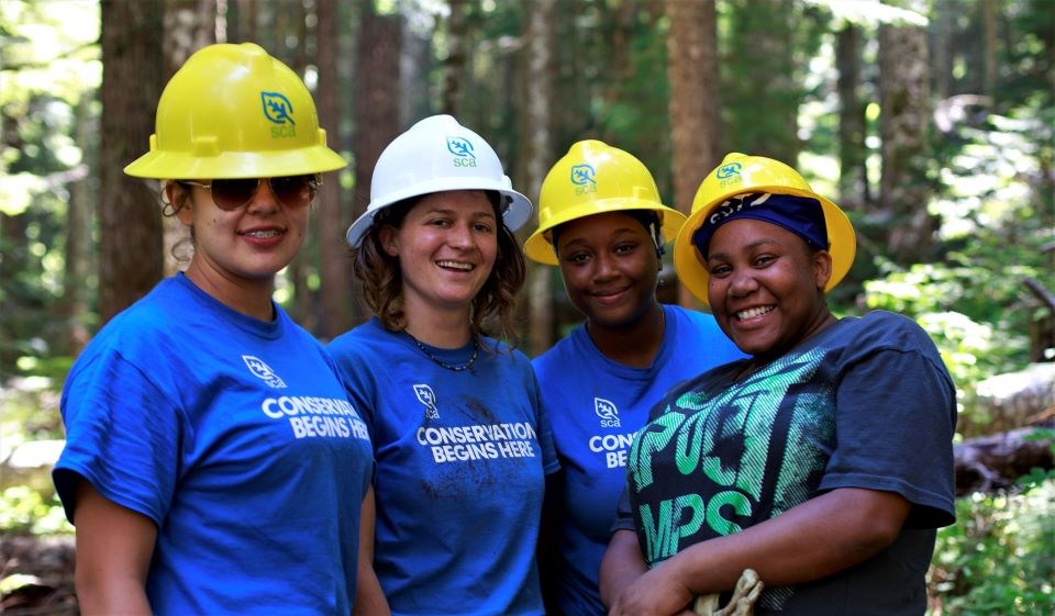 Four young adult volunteers in work gear smiling in the woods.