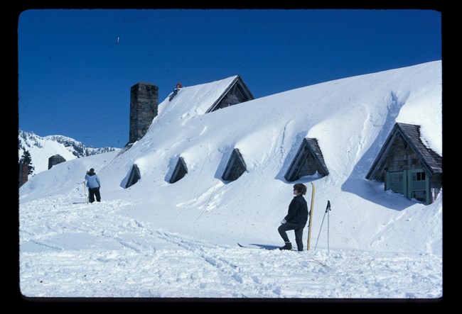 Skiers stand atop snow in front of second story windows of Paradise Inn.