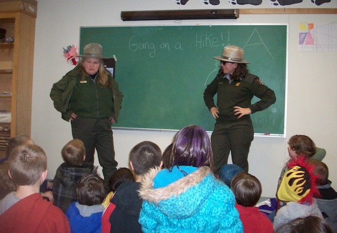 Two rangers speak to a classroom of elementary school children. They stand in front of a chalkboard with the text "Going for a Hike!" written on it. Two rangers speak to a classroom of elementary school children. They stand in front of a chalkboard with the text "Going for a Hike!" written on it.