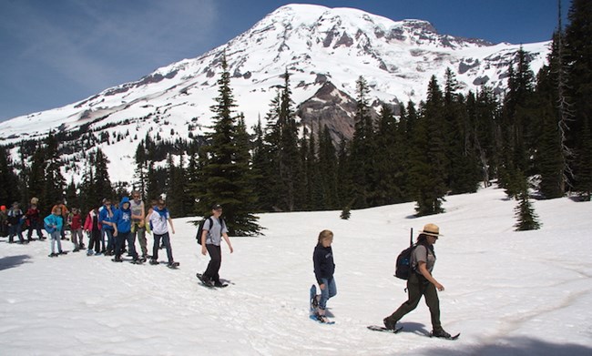 A ranger leads a group of students on snowshoes across a snow-covered meadow with views of Mount Rainier.