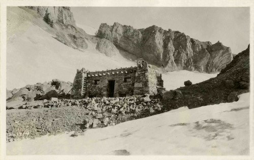 A rectangular stone building with a flat roof perched on a rocky ridge high on a mountain.