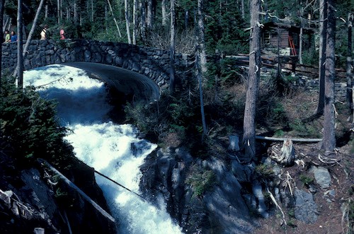 A stone-faced concrete bridge arches over a roaring white river. Several people cross the bridge. There is a small building on the right side of the bridge with a trail descending the slope to the right through the trees.