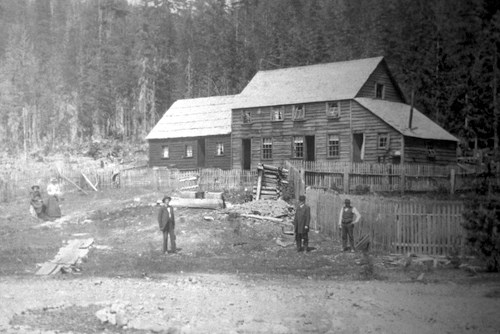 Black and white photo of a large wood building and several people standing in the dirt yard in front of it.