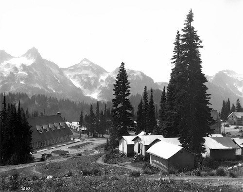 Black and white historic image of a cluster of tent cabins in a meadow next to a large wood building and a view of a rocky ridge.
