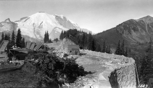 A rock retaining wall frames a road and parking lot under construction at the end of a ridge. Dump trucks are parked in the center, the backs tilted up. A glacier-covered mountain rises in the background.