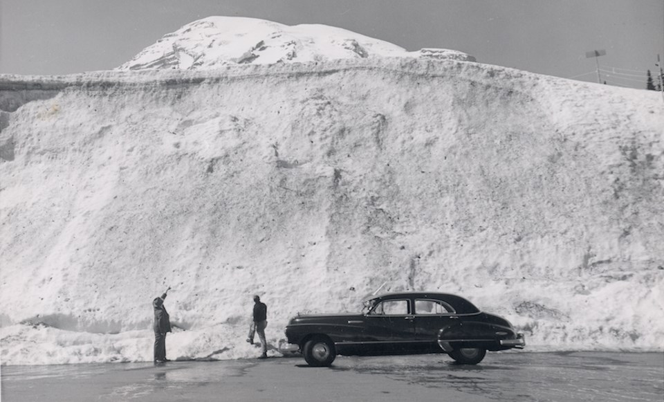 Black and white photo of two men standing next to an old car looking up at a huge snowbank.