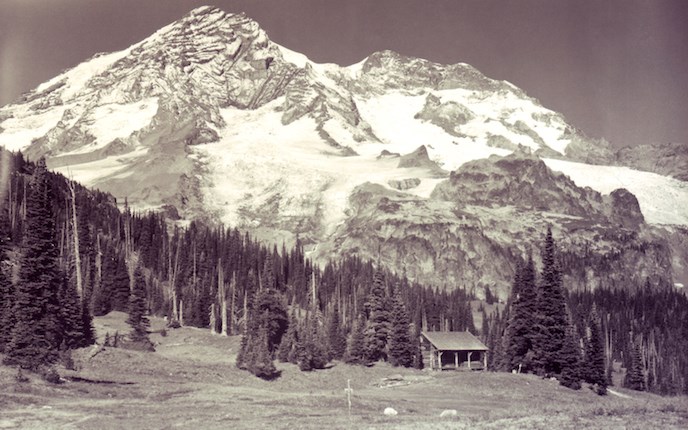 Black and white photo of Mount Rainier with tiny log cabin tucked amid the firs at the base of the mountain.