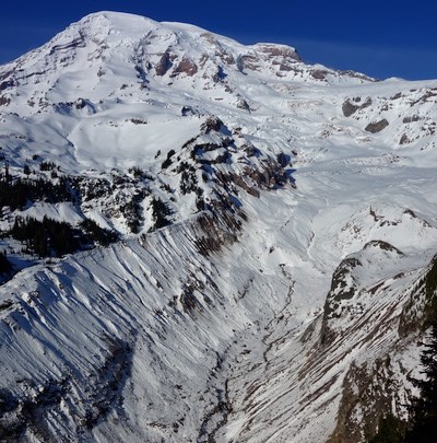 Nisqually glacier curves down the slope of Mount Rainier, a small creek emerging from the snow-covered terminus halfway up a deep valley banked with moraines.