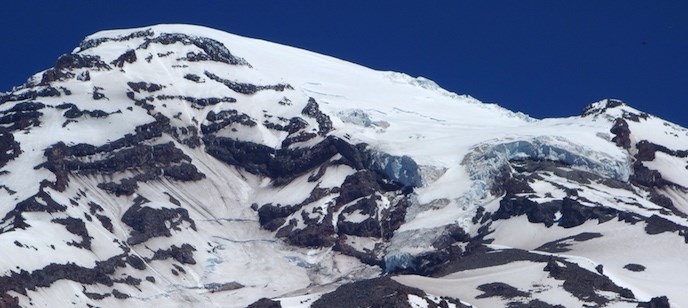 The summit of Mount Rainier with a thick glacier curling from the peak, and covered in patches of snow.