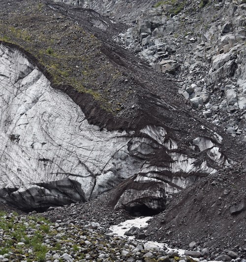 Dirt and rocks cover the ice terminus of a glacier, with a river emerging from a cave in the ice.