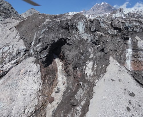 Aerial photo of a rocky glacier terminus with water pouring out of a cavern in the ice.