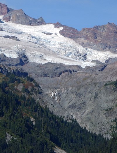 A distant waterfall flows over a cliff below a glacier and rocky ridge line.