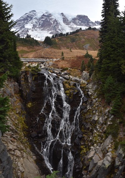 A braided waterfall flows over a cliff with Mount Rainier in the background.