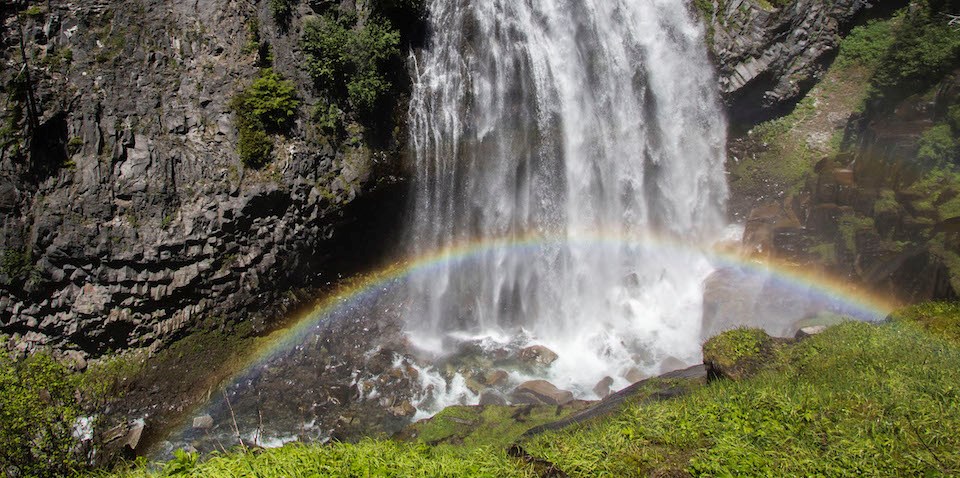 A rainbow arches through the mist at the base of a waterfall descending a rocky cliff.