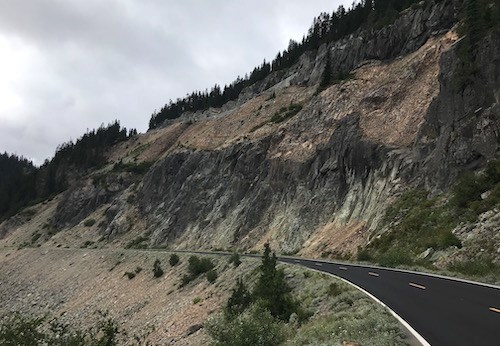 Thick bands of darker grey and crumbly tan-orange rock layered diagonally in a cliff face traversed by a paved road.