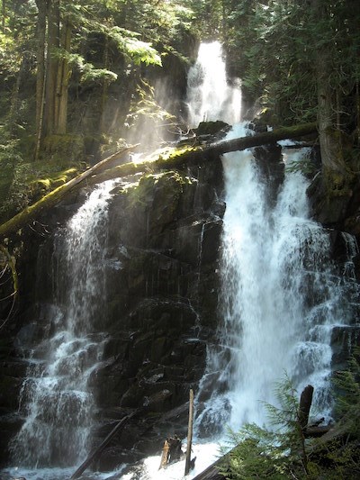 A waterfall spills around a rocky outcrop surrounded by dense forest broken by beams of sunlight.