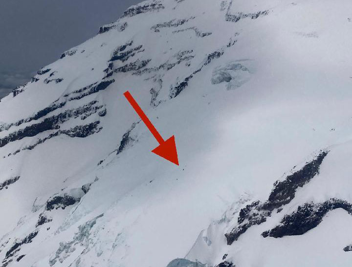 A red arrow points out a tiny dot on the slope of a glaciated mountain