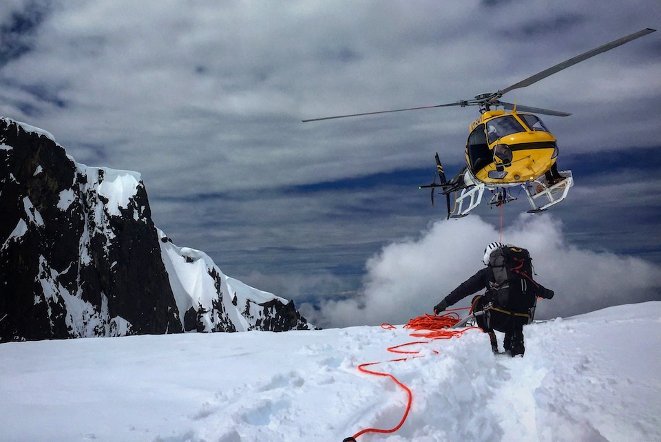 A person in a flight suit and helmet holding a red rope kneels in snow as a yellow helicopter approaches.