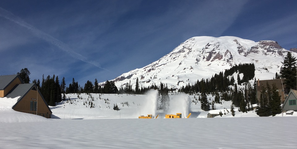 Two rotary plows blow plumes of snow up from a snow-covered parking lot on the slopes of Mount Rainier.