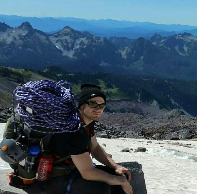A man with a large backpack with coils of rope sits on the edge of a snow field overlooking a view of mountain ranges.
