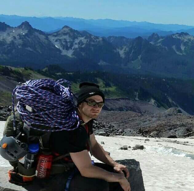 Alex Fitzgerald A man with a large backpack with coils of rope sits on the edge of a snow field overlooking a view of mountain ranges.