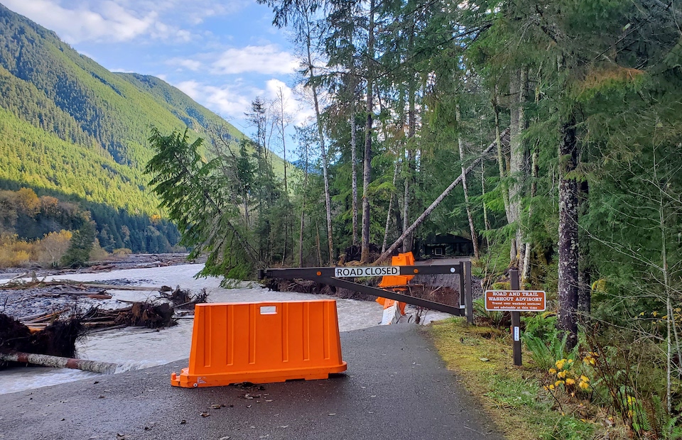 An orange barrier and half of a metal gate block access to a former road that has collapsed into a river.