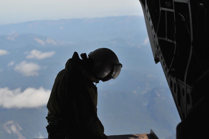 A silhouette of a person wearing a flight helmet with head bowed looks out the open door of a helicopter.