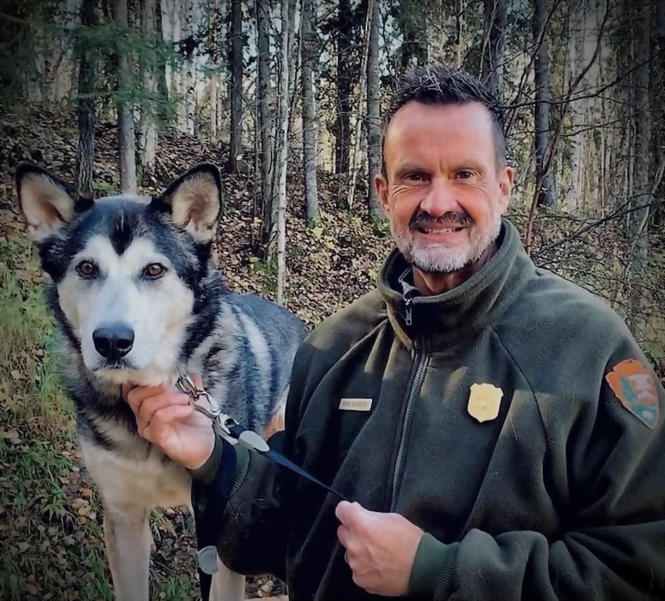 A person in a green National Park Service Uniform stands next to a black and white dog.