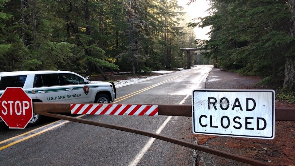 A "road closed" and stop sign attached to a metal gate that blocks a two-lane paved road with a US Park Ranger vehicle parked behind the gate. A wooden entrance arch frames the road a short distance away.