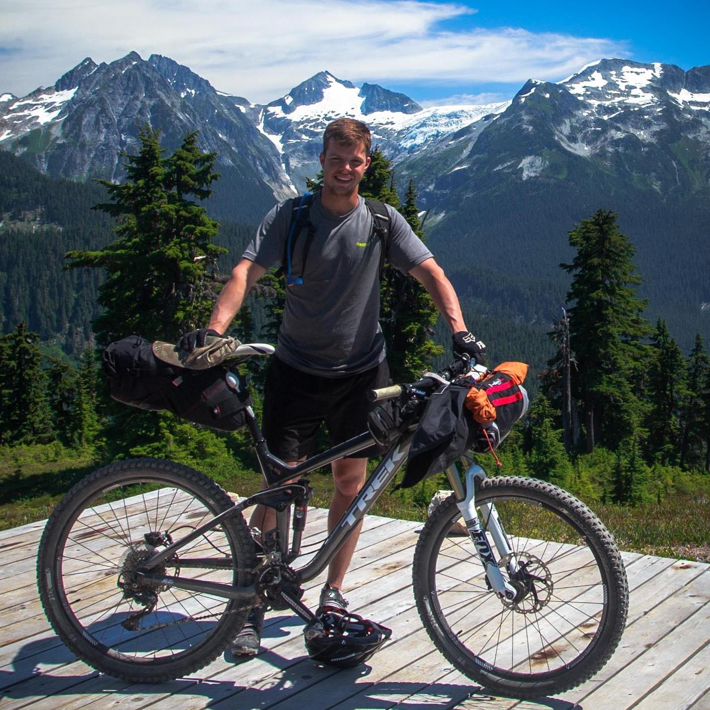 Matthew Bunker Matthew Bunker stands with his bicycle with snowcapped mountains in the background.