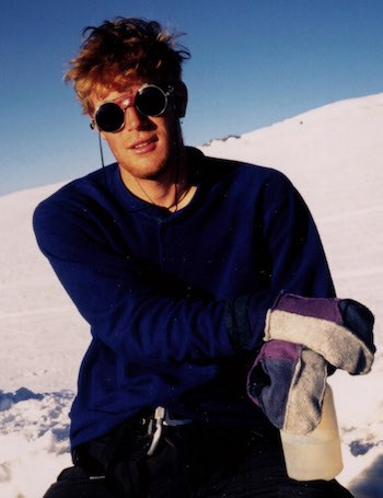 A man kneels on a snow slope, hands in gloves crossed over one knee.