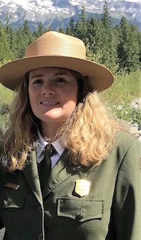 A woman with long blonde hair in an NPS uniform in front of trees