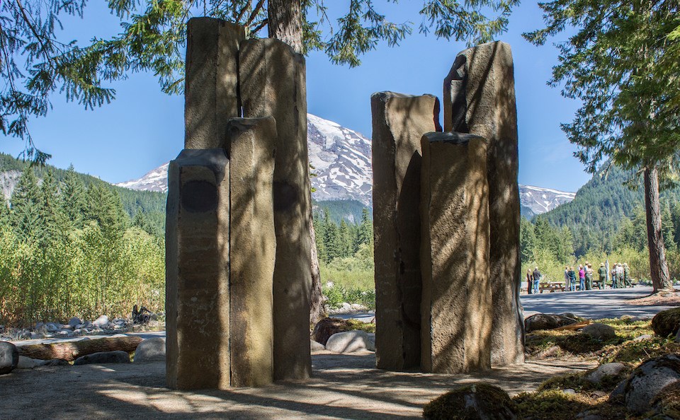 Two clusters of tall basalt columns in front of a view of Mount Rainier, shaded by trees.