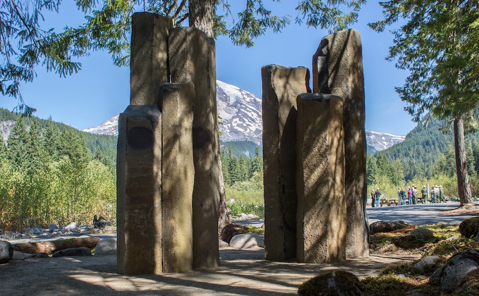 Two clusters of tall basalt columns in front of a view of Mount Rainier, shaded by trees. Two clusters of tall basalt columns in front of a view of Mount Rainier, shaded by trees.