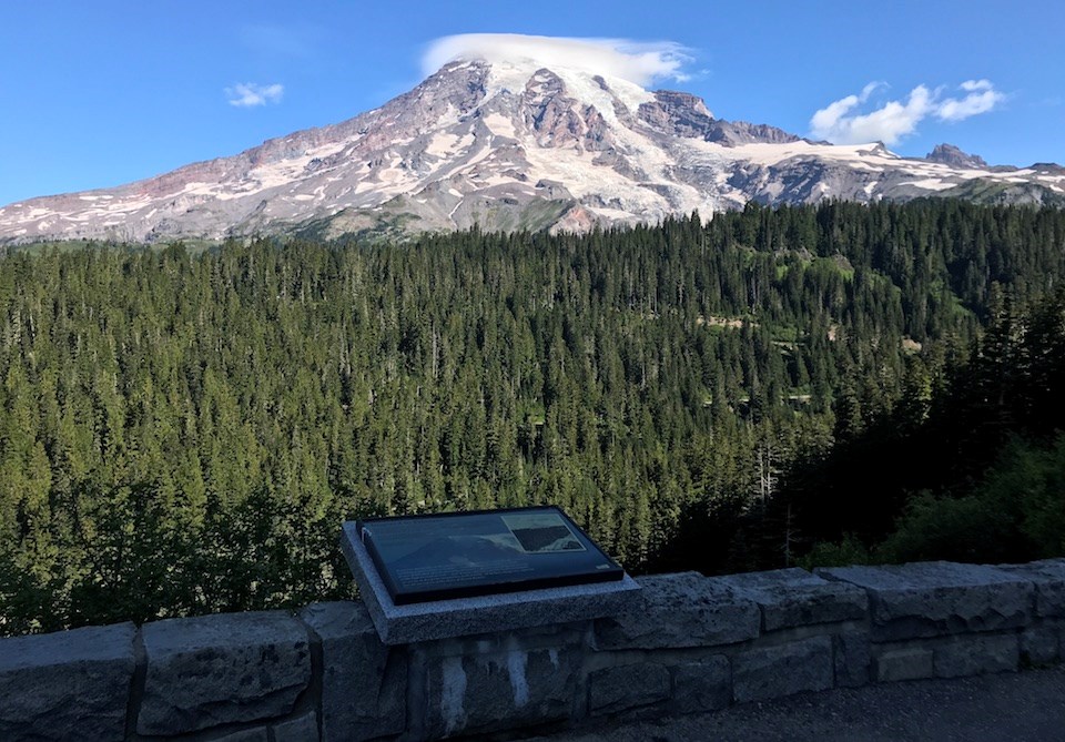 A wayside panel sits on a low rock wall in front of a view of a forested slopes that give way to a glaciated mountain peak capped in clouds.