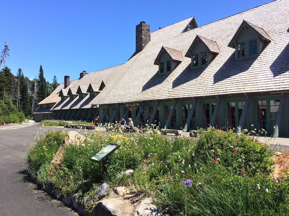 A small wayside stands in an island of flowers in a parking lot in front of a large wood building with a steeply-angled roof.