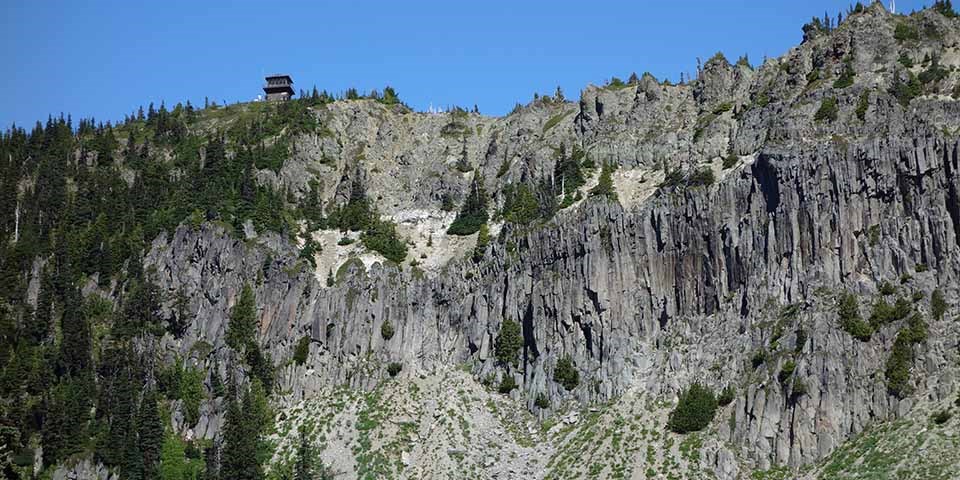 A small lookout cabin perches on a ridge line above a rocky cliff.