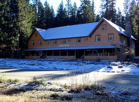 A wood building with two gabled ends and a wrap around covered porch frosted by snow and surrounded by forest.