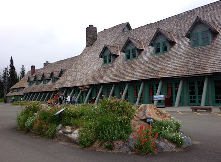 A long building with a steeply angled wood shingle roof, gabled windows and wood beams covering rows of windows on the ground floor. Wildflowers bloom in an island in the road in front of the building.