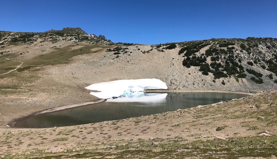 A small lake on rocky slope with sparse patches of meadow vegetation. A small snowbank clings to one edge of the lake.