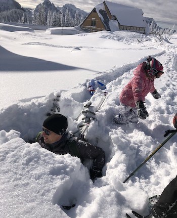 A man up to his neck in snow next to two small trees while a small child stands next to him on top of the snow.