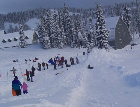 Brightly dressed childern and adults playing in the snow in front of rustic historic buildings at Paradise.