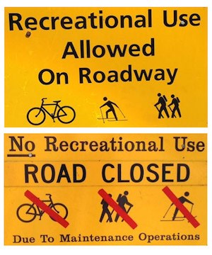 Two yellow road signs, text reads (top) "Recreational Use Allowed on Roadway" with symbols of a bicycle, skier, and hikers. Bottom text reads "No Recreational Use Road Closed due to Maintenance Operations" with symbols of bicycle, hikers, and skier.