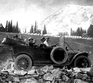 Visitors sit in an open-top old car in front of Mount Rainier in a black and white photo.