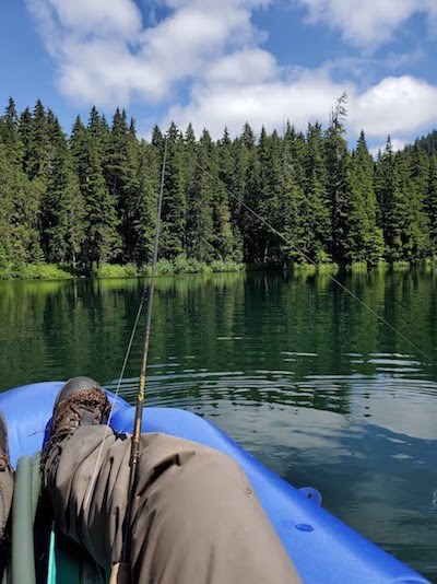 First-person view of a person fishing from a boat, fishing rod propped up on their knee, from a boat in a still lake surrounded by forest.