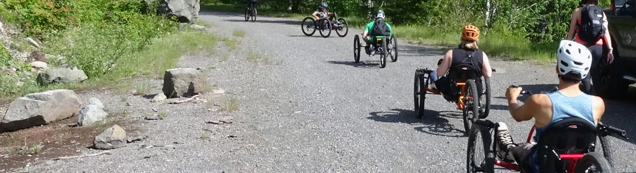 Handcyclists on the Westside Road A group of handcyclists travel up a gravel road surrounded by tall evergreen trees.