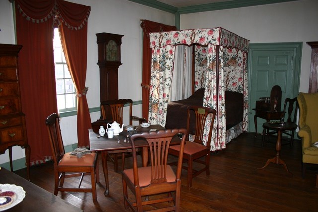 A view of a well furnished early American bedroom, with wooden chairs, a four poster bed, and a tall case clock among the objects displayed.