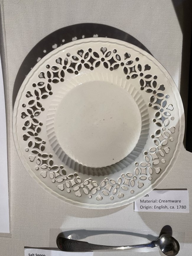 A dish made of creamware with a lacy design around its edge made with holes going through the dish itself, on display above a long silver salt spoon in a museum case.