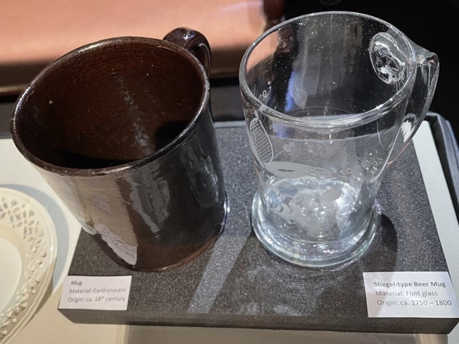 A brown earthenware mug and a glass stein vessel with handle, side by side on a pedestal in a glass museum case.